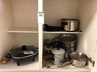 Metal pots and pans arranged on white shelving including Rival electric skillet on bottom shelf with glass lid and various aluminum and stainless steel pots and pans above.