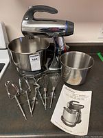 Full view of Sunbeam Mixmaster stand mixer with two stainless steel bowls, six accessory attachments (two beaters, two dough hooks, two whisks), and the user manual spread out on countertop.