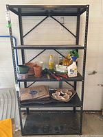 Front view of black metal utility shelf with five shelves holding gardening and household items, against a garage wall. Shelf shows wear and tear.