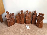 Six ornate wood carvings depicting abstract human pairs, standing on carpet against a wall. Signed pieces, polished with natural wood grain visible.