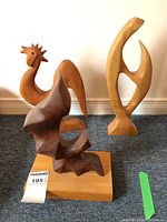 Three wooden carvings placed on dark carpeted floor against white wall, includes light wood rooster carving, dark wood abstract sculpture on wooden base, and light wood abstract sculpture.