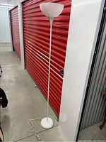Full view of the white metal floor lamp next to a red shutter door, showing its height and design.