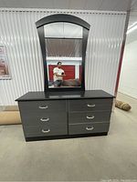 Front view of the dresser with attached mirror in a storage space, showing six drawers and a large rectangular mirror with rounded top.