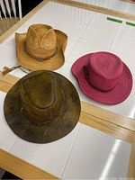 Top view of three hats on a white table showing brown leather cowboy hat, tan straw cowboy hat, and pink wide-brimmed hat