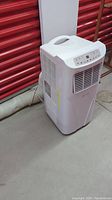 White Kross portable air conditioner seen front and side with attached hose on floor.