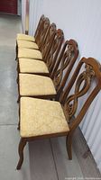 Six wooden dining chairs aligned in a row showing carved wooden backrests and golden yellow patterned seat cushions.