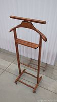Front view of vintage solid wood valet stand showing hanger, tray, and base with wheels on concrete floor.