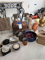 Various copper and brass items arranged on carpet including pitchers, metal cups, coasters, a carved stone plaque, a bowl of red glass stones, and decorative metal box.