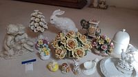 Overview of porcelain and ceramic decorative items showing figurines of children, rabbits, floral arrangements, and a white lidded container.