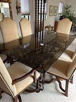 Dinning table with smoked walnut wood top and ornate wrought iron base surrounded by six beige upholstered chairs with wooden legs and nailhead trim.
