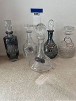 Front view showing five etched glass decanters, two smoked glass and three clear glass, with various shaped stoppers and metallic labels hanging on chains.