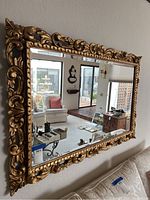 Large rectangular mirror mounted on a wall in a living room, showing gold ornate frame with carved scrollwork and leaf patterns, glass beveled at edges.