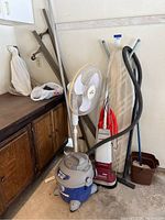 Overview of ironing board, standing fan, Shop Vac, shampoo polisher, mop, bucket, and bed frames against wall near cabinet.