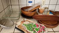 Photo showing assortment of kitchen items on counter including two clear rectangular Pyrex dishes, wooden bowls and salad utensils wrapped in plastic, long oval wooden serving board, various smaller wooden boards, ceramic bowl with radish painting, ceramic pot, and mesh strainer with handle.