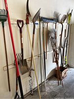 Full view of assorted gardening tools including rakes, shovels, hoes, scythe, and push broom hanging on wall