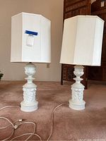 Two white porcelain lamps with cherubs standing on a carpeted floor, hexagonal white fabric shades on each lamp.