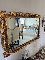 Full view of the gold painted ornate wall mirror hanging on a wall, reflecting chairs and room interior.