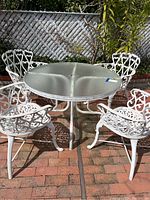 Full view of the round glass patio table with four white wrought iron chairs set on a brick surface outdoors in front of a lattice fence.