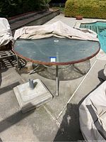 Large round metal patio table with glass top and cement umbrella stand on concrete patio.