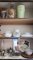 Wide view of shelves with multiple white ceramic serving dishes stacked along with some small utensil holders and decorative paper items at bottom shelf.