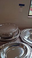Various silverplate serving trays and dishes stacked and leaning against a wall showing different sizes and embossed decorative patterns.