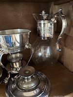 Glass pitcher with decorative silver-plated handle and lid, silver-plated chafing dish stand with inner bowl, silver-plated covered serving dish on wooden shelf.