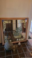 Photo shows large square mirror with wooden frame featuring southwest style geometric patterns at corners and central cactus design. Two pottery pieces and two small ceramic items placed in front.