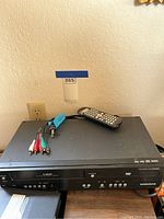 Magnavox VHS player with remote and AV cables on top, set against a plain wall