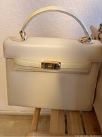 Structured cream-colored handbag placed on a bamboo bench showing front side with clasp and handle.