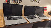 Three silver Apple MacBook Pro laptops side by side on a wooden surface, screen off, keyboard visible, all showing signs of wear.