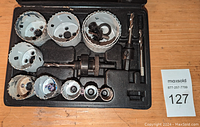Photo showing multiple hole saws of varying sizes, arbors, drill bits, and screws inside a black molded carrying case on a wooden surface.