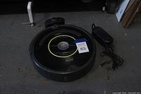 Top and angled view of the black iRobot Roomba robotic vacuum with yellow ring around central button, placed on a floor with charger and power cord beside it.