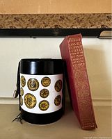 Front and side view of the vintage money box featuring various coin illustrations on a white band wrapped around a black metal cylinder.