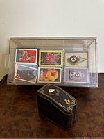 Photo showing the complete vintage Lionna black leather playing card box with gold decoration and a clear plastic case holding six decks of playing cards each with unique cover designs including cats, flowers, clocks, and Hawaii theme.