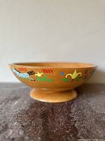 Side view of the wooden bowl showing colorful hand-painted decorations of animals and flowers on the side of the bowl, natural wood finish, footed base.