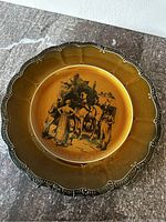 Photo of the front of the Ridgway vintage plate depicting coaching scene with figures and horses, detailed black transfer illustration on brown amber glaze plate with scalloped rim.
