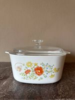 Front view of vintage white opaque glass CorningWare casserole dish with colorful Wildflower pattern and clear glass lid with knob handle on top.