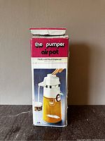Original box of 'the pumper' airpot liquid dispenser with colorful red, white, and yellow design and photo of carafe.