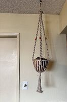 Full view of the hanging pot suspended from the ceiling showing the beige macramé hanger with red beads and braided tassel at the bottom.
