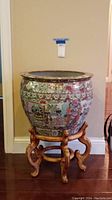 Full view of large round Asian-style ceramic planter with detailed colorful scenes and wooden carved stand.
