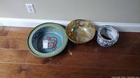 Front view of three items showing the large blue dragon bowl, gold floral bowl, and white and blue planter arranged on wooden floor.