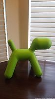 Side view of a green puppy shaped plastic figure on wooden surface near window blinds.