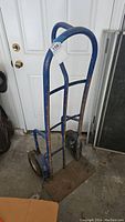 Front view of a blue metal rolling hand truck with rust and wear on the frame and wheels, against a white door background.