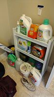 Full view of white plastic shelving unit holding household cleaning supplies including Tide detergent, Clorox, Bounce, spray bottles, bucket, and Pitch-It IV Pole.