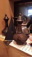 Three metal items displayed on a shelf including the metal kettle, textured coffee pot, and black pyramid decorative item.