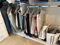 Closet rod with multiple men's pants and shirts hung, some clothing items in garment bags and plastic covers, showing variety of casual and golf pants