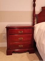 Full view of the reddish-brown wooden nightstand against a beige wall next to a bed frame with a carved post.