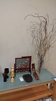 Overall display of all lot items on a glass table against a wall, showing clear tall glass vase with decorative branches, embroidered bird framed art, figurine, and wood decorative pieces.