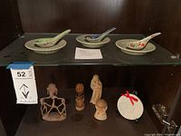 Photo showing three Asian vintage ceramic wonton spoons each paired with a matching saucer on glass shelf, above six assorted figurines including metal, wooden and ceramic pieces.