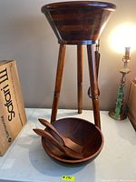 Floor standing wooden bowl stand with large wooden bowl on top and small wooden bowl below, with two wooden salad utensils placed inside the small bowl